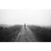 A black and white photograph of a person standing on a dirt path in a foggy outdoor landscape with tall grass on both sides.