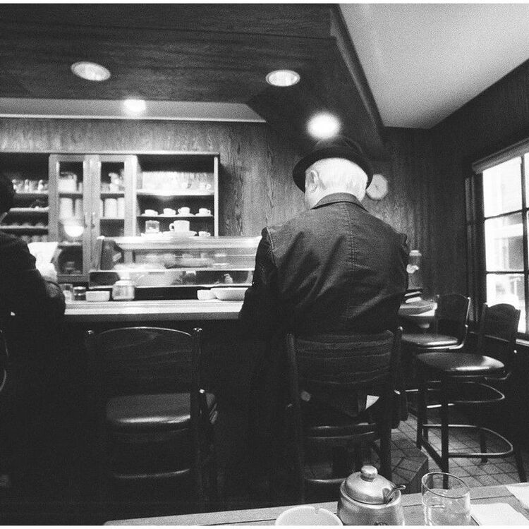 Black and white photograph of a man sitting at a diner counter, viewed from behind as he looks toward the kitchen display case.
