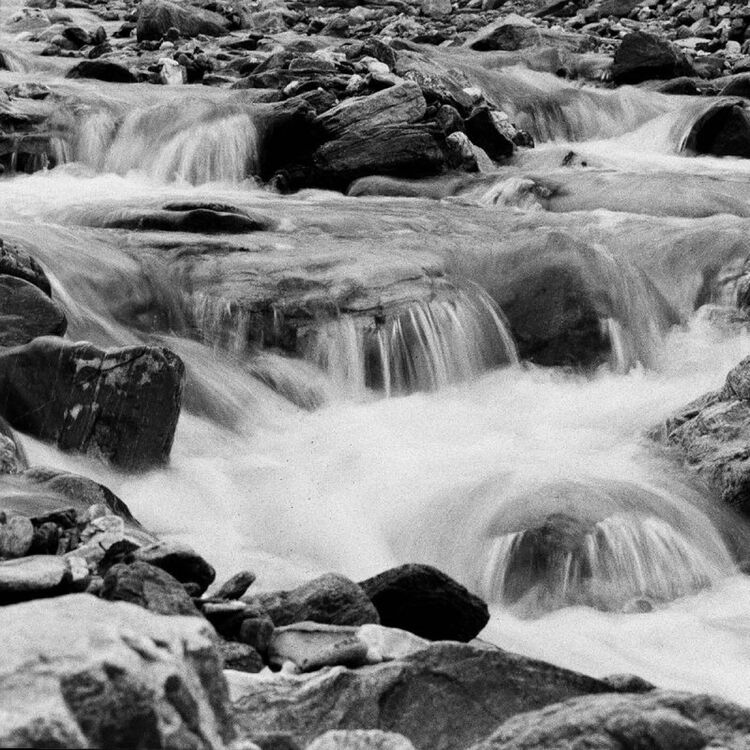 Black and white photograph of a rocky stream with multiple small cascades flowing over stones, creating white water throughout the frame.