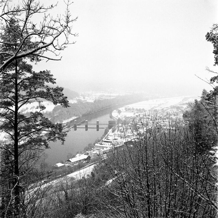 Black and white landscape photograph of a snowy river valley with a bridge spanning the water, surrounded by bare winter trees and a small town in the distance.