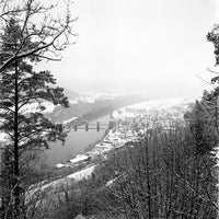 Black and white landscape photograph of a snowy river valley with a bridge spanning the water, surrounded by bare winter trees and a small town in the distance.