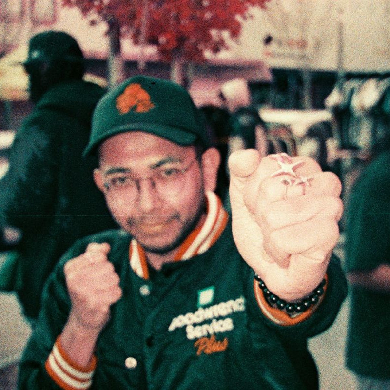 Person wearing a green jacket and cap, holding a small object in an urban setting.