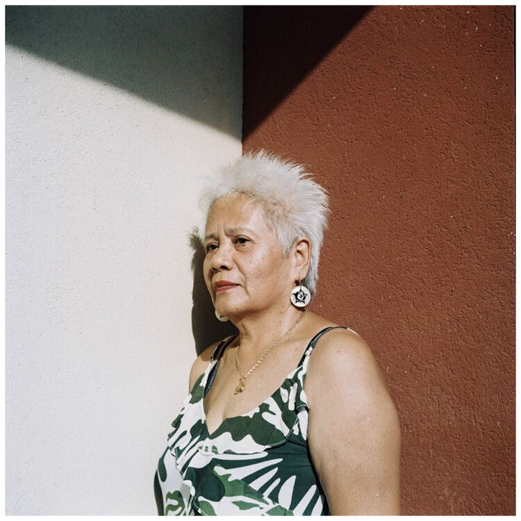 An older woman with short white hair stands against a split white and reddish-brown wall, wearing a green and white patterned sleeveless top and large round earrings.
