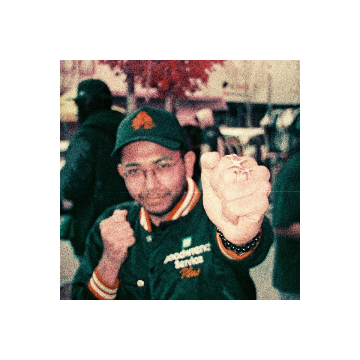 Person wearing a green jacket and cap, holding a small object in an urban setting.