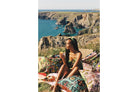 Woman sitting on a picnic blanket with patterned cushions overlooking a sunny coastal cliffside in vibrant film colors.