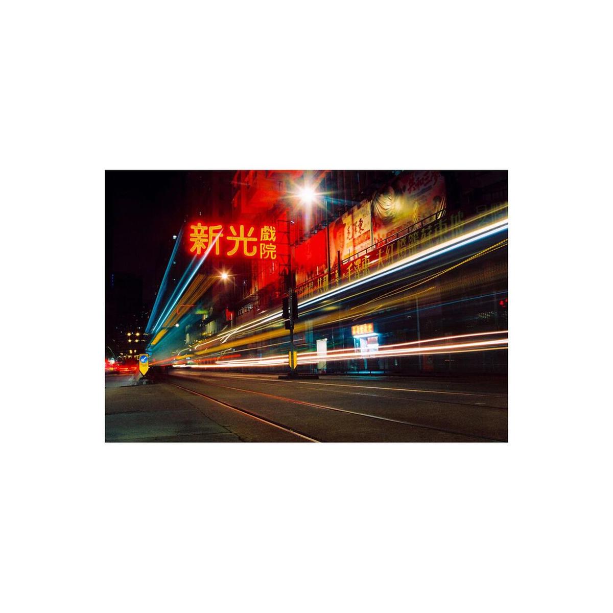 A long-exposure city street scene at night with bright red and yellow neon signage, blurred vehicle light trails, and a dark urban background.