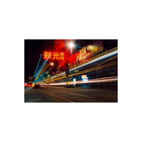 A long-exposure city street scene at night with bright red and yellow neon signage, blurred vehicle light trails, and a dark urban background.