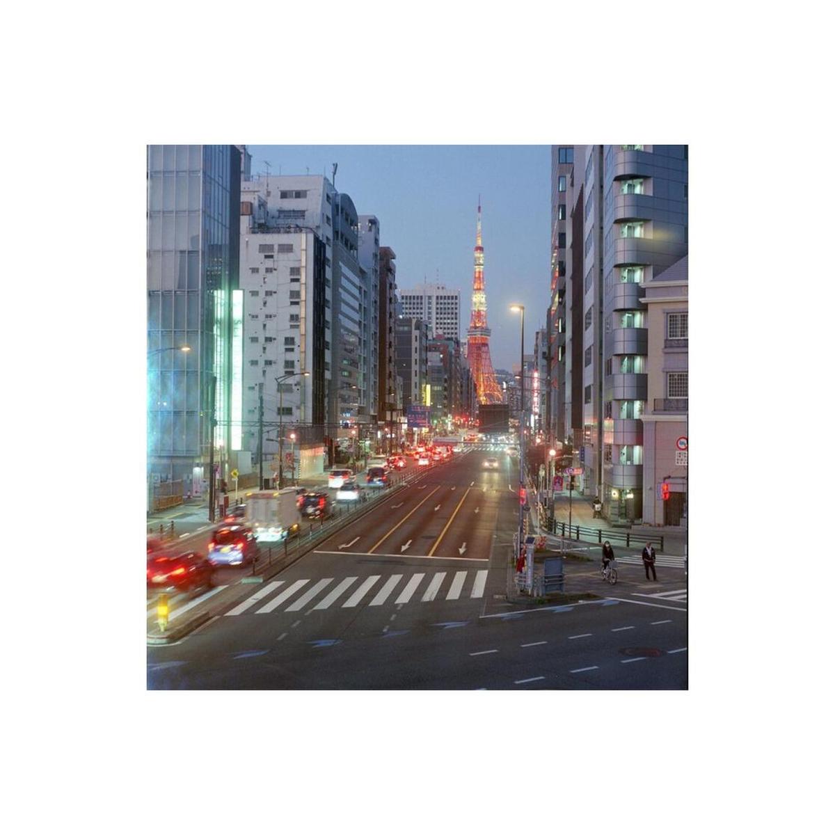 A scenic urban street in Tokyo at dusk, with tall buildings on both sides, cars with illuminated taillights on the road, and the Tokyo Tower visible in the distance against a blue evening sky.