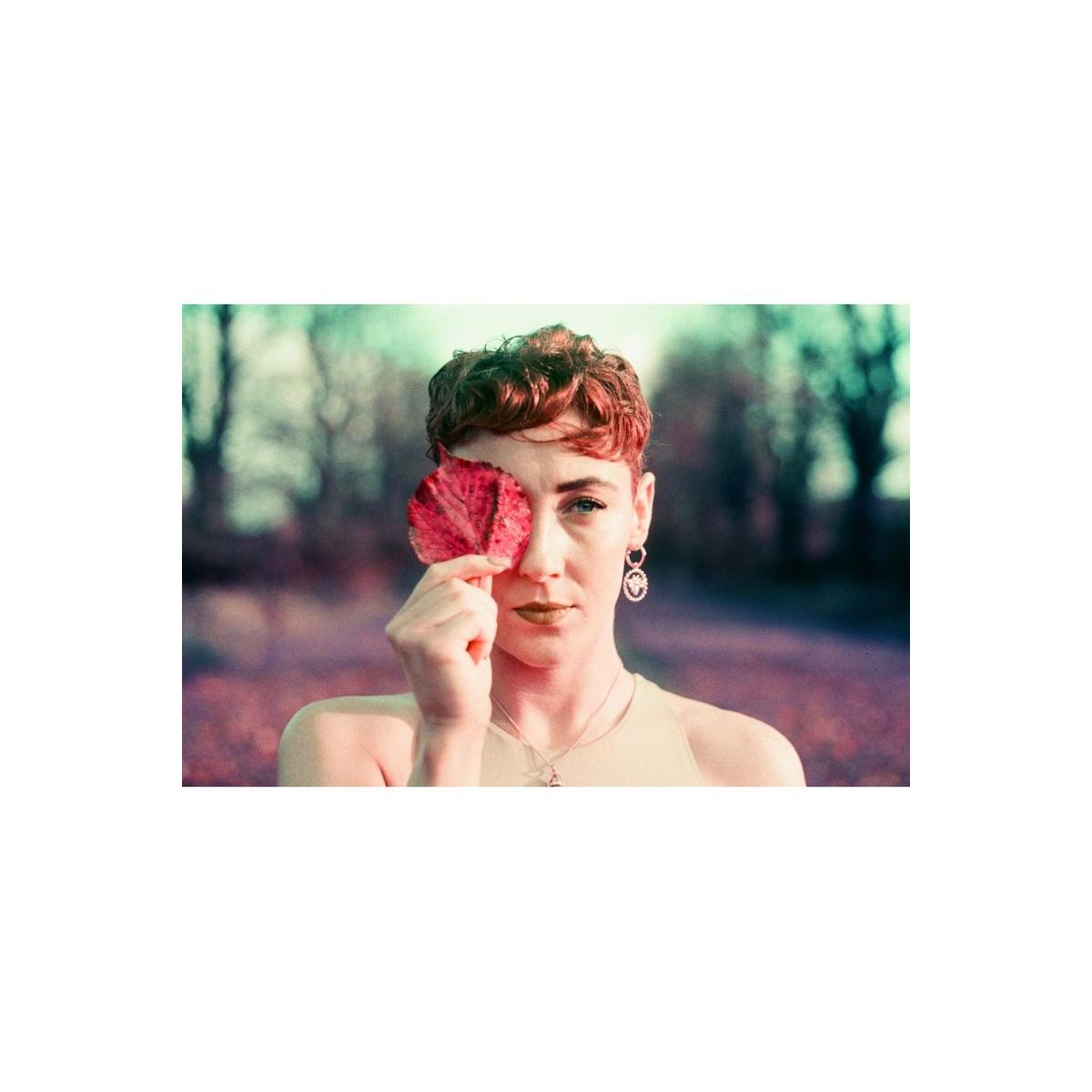 Person holding a red leaf over one eye, captured with vintage film tones and purple color shifts.