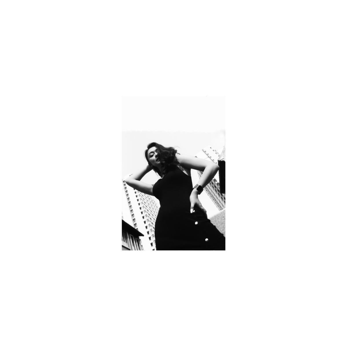 Low-angle black and white portrait of a woman in a dark dress against city buildings, captured on Babylon Kino 13 film.