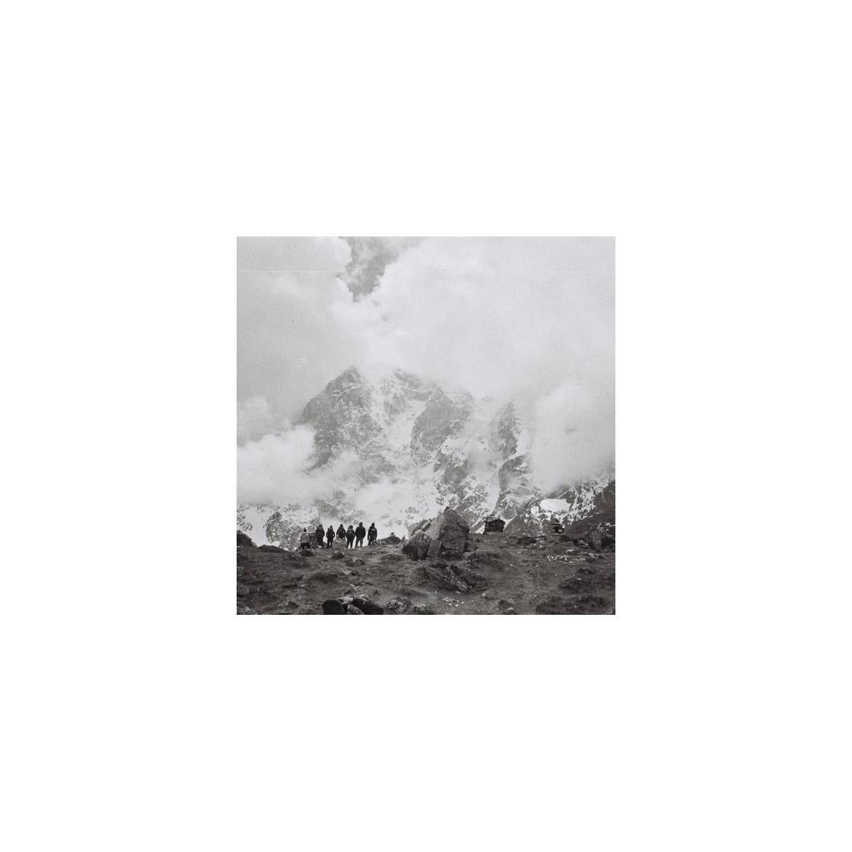 Black and white square-format film shot of a group of hikers standing before a cloudy, snow-capped mountain peak.