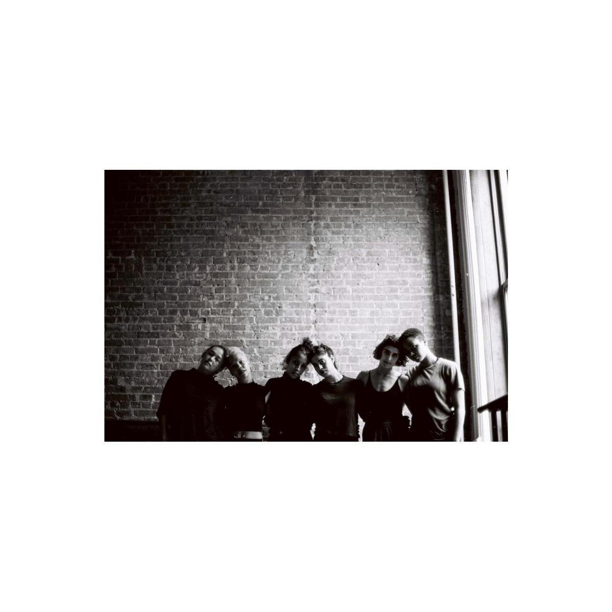 Six people standing in a row against a brick wall in a high-contrast black and white film portrait.