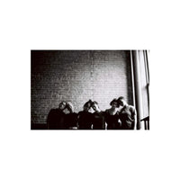 Six people standing in a row against a brick wall in a high-contrast black and white film portrait.