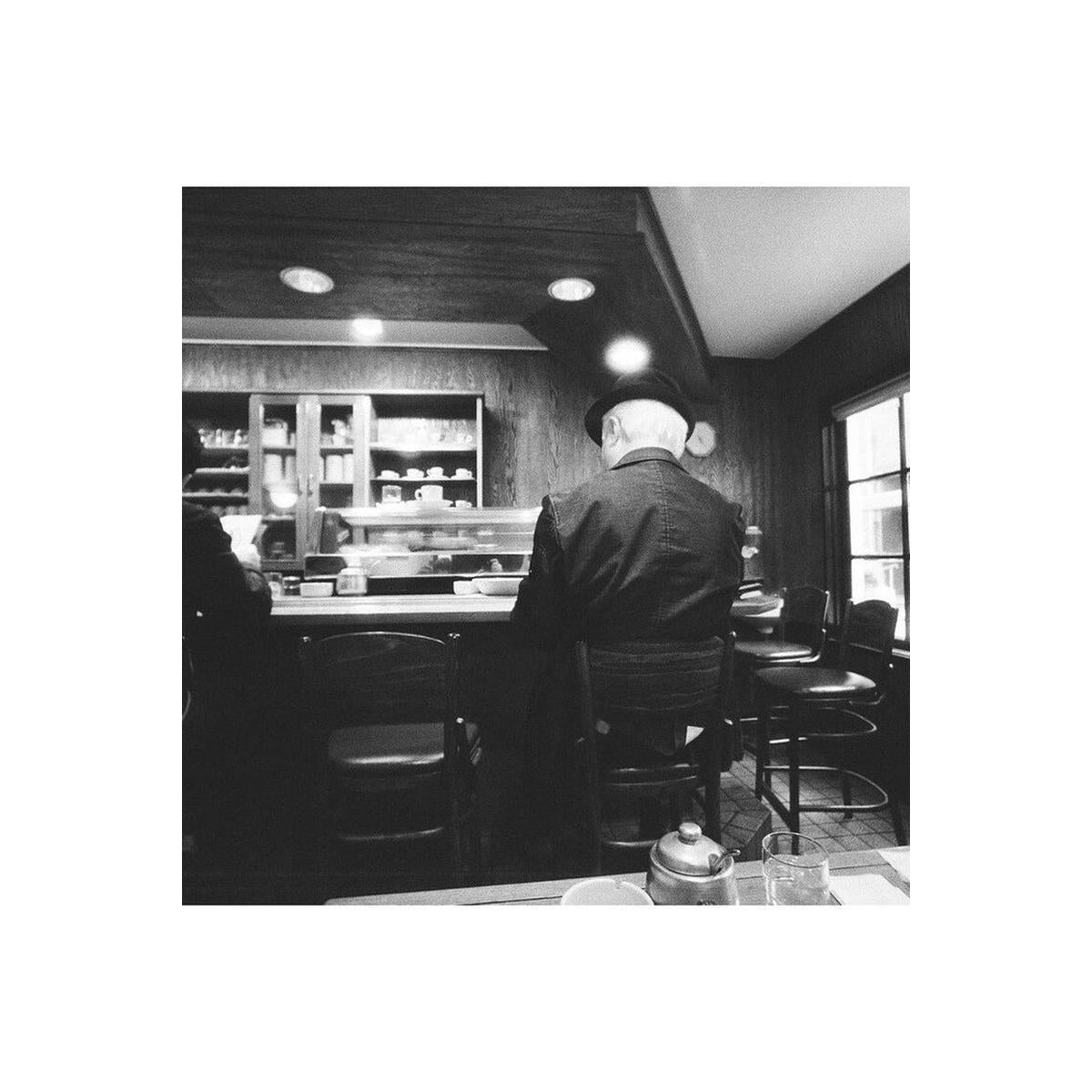 Black and white photograph of a man sitting at a diner counter, viewed from behind as he looks toward the kitchen display case.