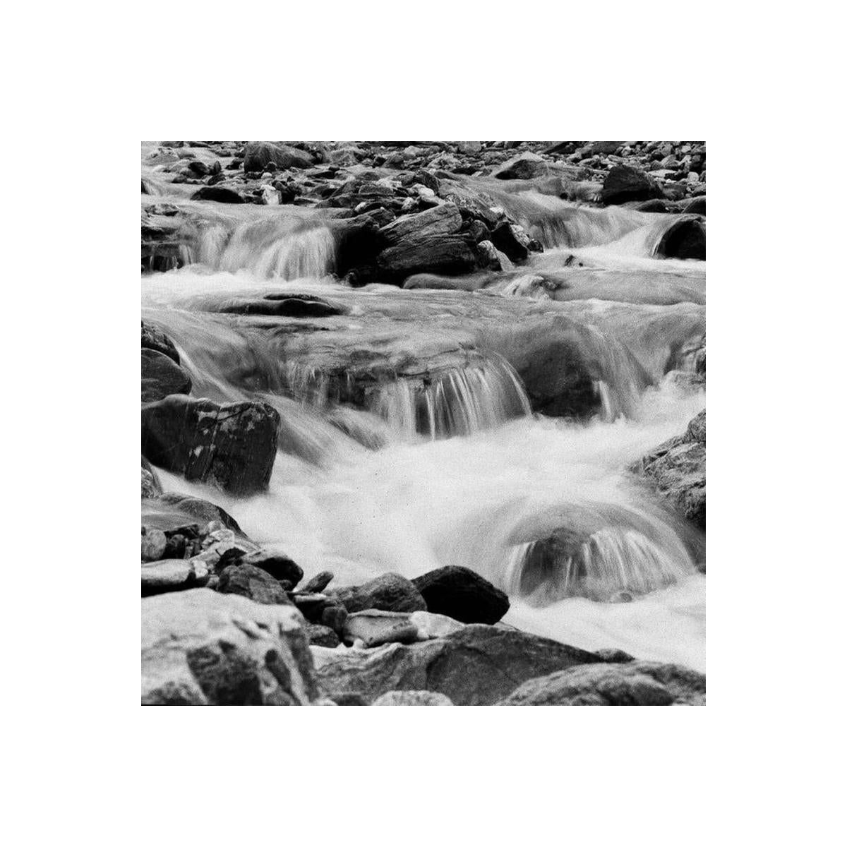 Black and white photograph of a rocky stream with multiple small cascades flowing over stones, creating white water throughout the frame.