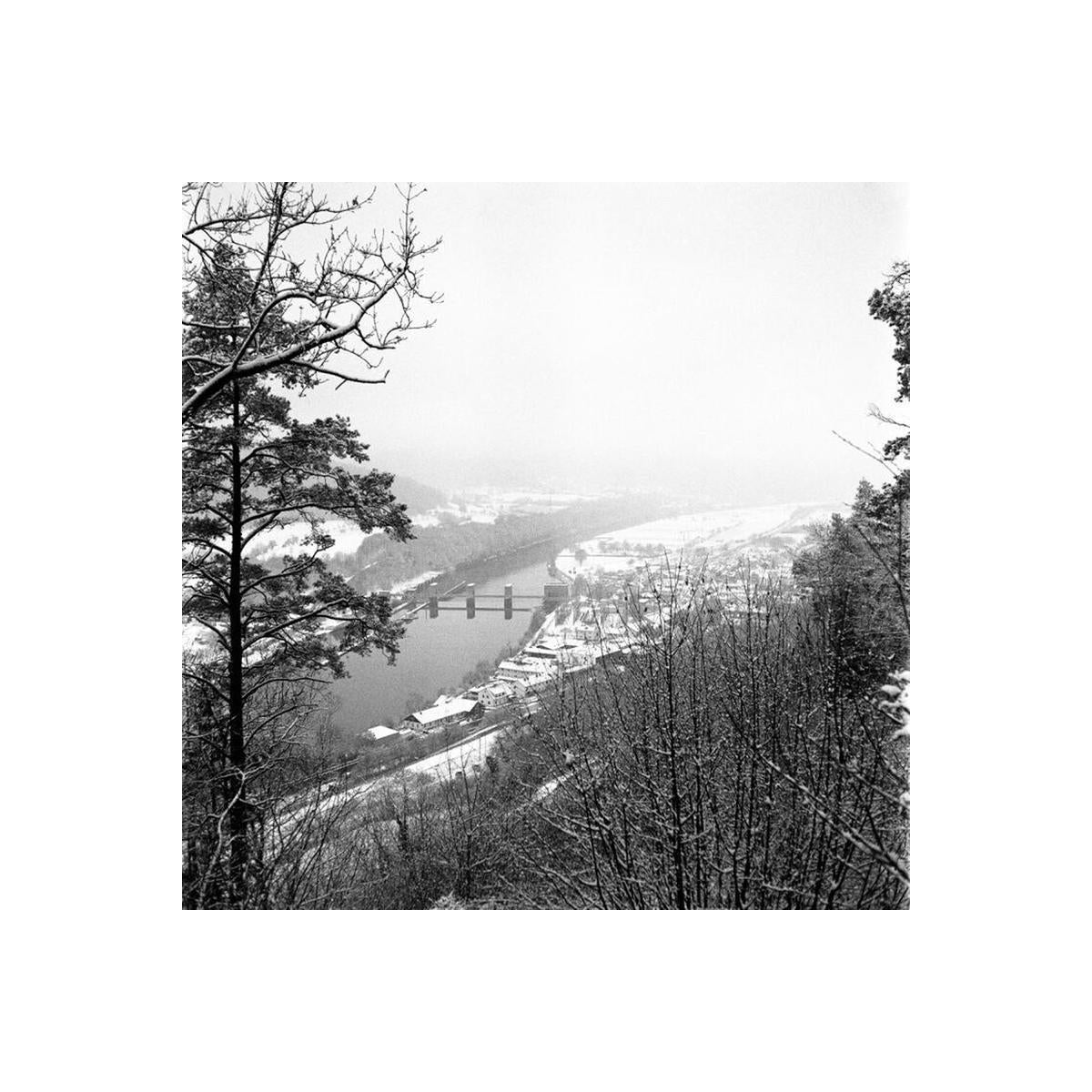 Black and white landscape photograph of a snowy river valley with a bridge spanning the water, surrounded by bare winter trees and a small town in the distance.