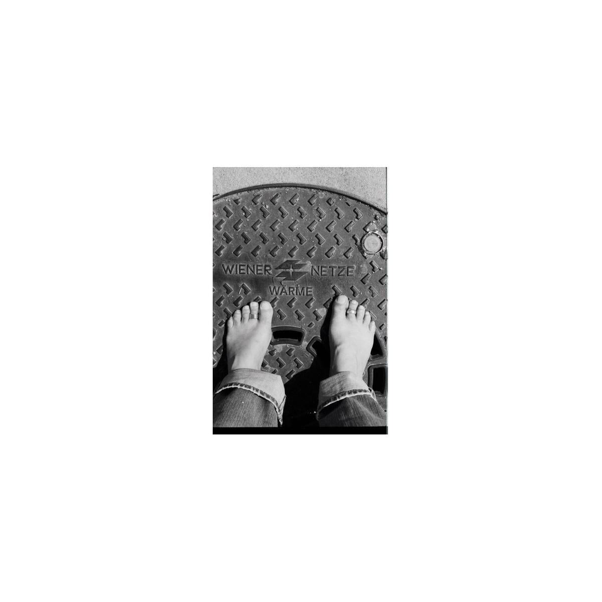 Bare feet standing on a textured metal manhole cover, captured in high-contrast black and white 35mm film.
