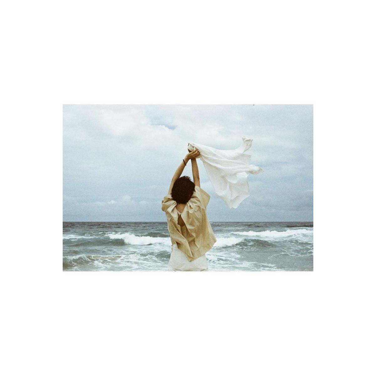 Person standing on a beach holding a white cloth, captured with the muted, high-contrast aesthetic of Lomography Metropolis.