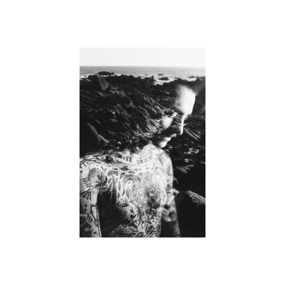 Double exposure black and white portrait of a tattooed man overlaid with a rocky coastal landscape.