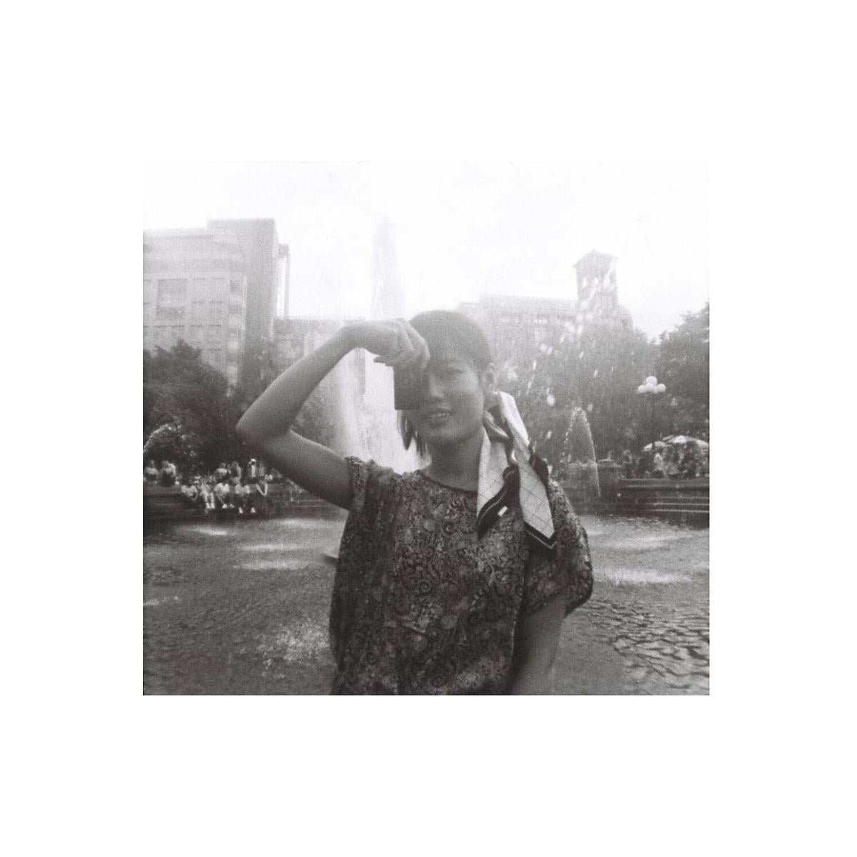 Person holding a small 110 camera in a park, captured in high-contrast black and white film with a grainy, vintage aesthetic.