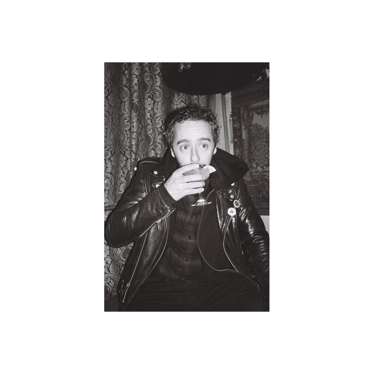 A black and white portrait of a man wearing a shiny leather jacket, holding a glass near his mouth, with patterned curtains and a dark hat in the background.