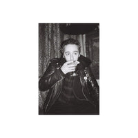 A black and white portrait of a man wearing a shiny leather jacket, holding a glass near his mouth, with patterned curtains and a dark hat in the background.