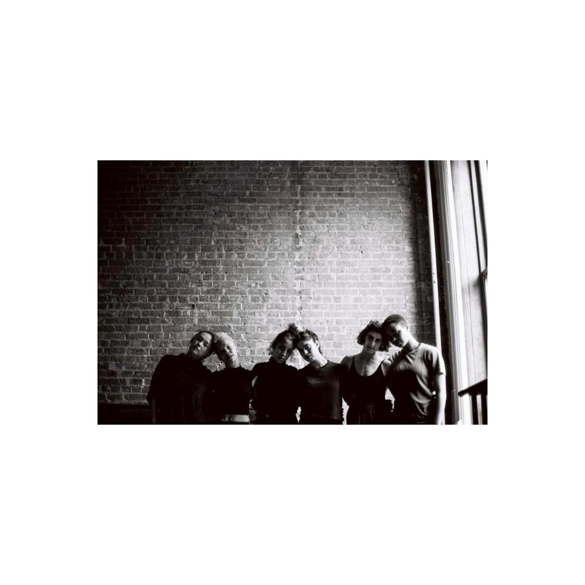 Black and white photograph of five people standing together against a brick wall, with a window light shining in from the right side.
