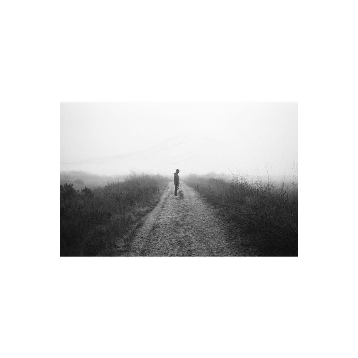 Person standing on a foggy dirt path in a high-contrast, black and white cinematic landscape shot.