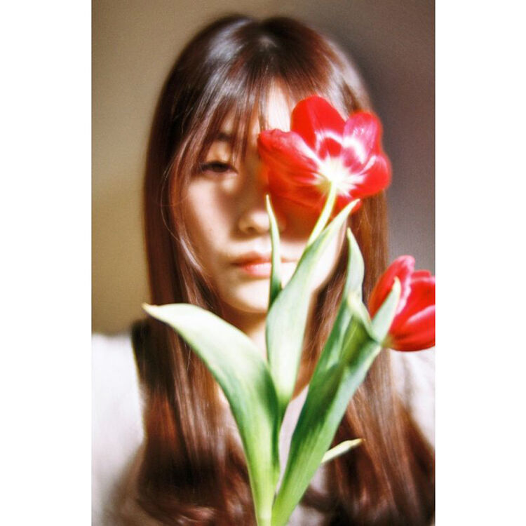 A portrait of a young woman with long brown hair holding two red tulips in front of her face, with a soft beige background.