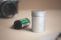 Roll of film with a green label next to tin container that says LOMO Film with a blurred camera in the background.