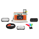 A retro-style Polaroid camera with a textured orange body, cream top, and black lens, shown with multiple accessories including a close-up lens, color filter, kiosk lens, strap, and color slides on a white background.