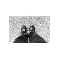 A top-down view of a person's legs in dark jeans and black lace-up shoes standing on a textured gray asphalt surface.