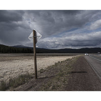 A rural landscape scene with a weathered diamond-shaped road sign on a wooden post beside a dirt road next to a grassy field, under a dramatic cloudy sky with distant mountains and a small vehicle on the road.
