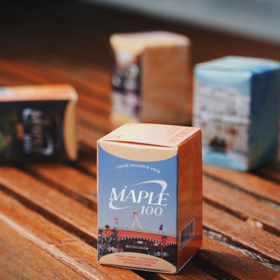 A boxed film canister labeled MAPLE 100 stands on a wooden table, with other blurred boxes in the background.