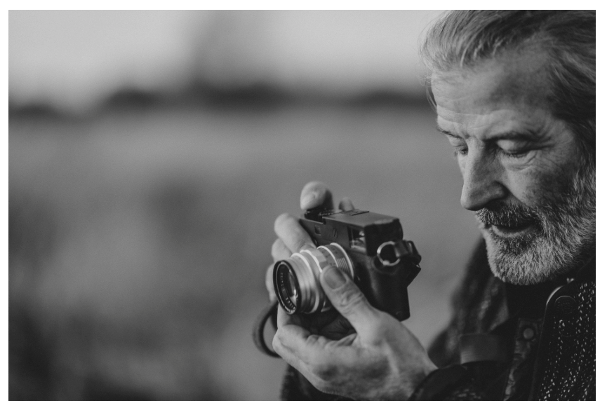 A black and white close-up photograph of an older man holding a vintage film camera, looking down at the lens with a focused expression.