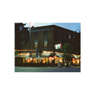 A square photographic print depicting a brick commercial building at dusk, with lit windows, awnings, outdoor dining umbrellas, and warm string lights along the front, centered on a white background.