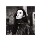 A black and white portrait of a woman with long dark hair wearing a shiny dark jacket, shown against a blurred urban background with a white border around the square photo.