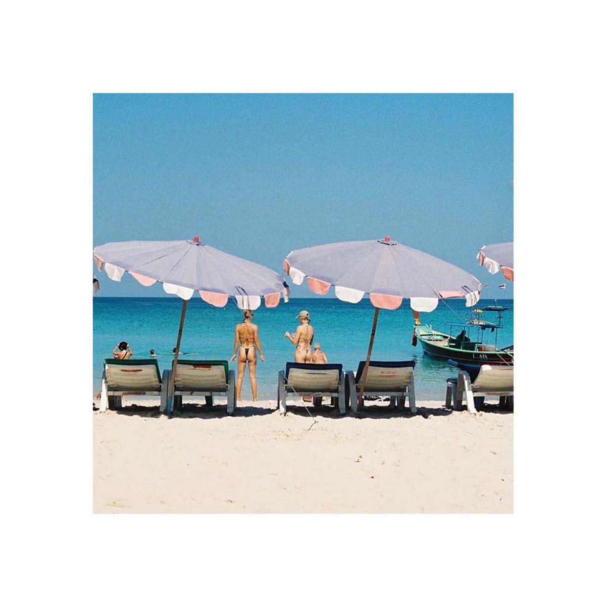 A square beach scene print showing white umbrellas over lounge chairs on a sandy shore, with turquoise water and a clear blue sky. Two people are visible near the umbrellas, and a small boat appears on the right.