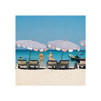 A square beach scene print showing white umbrellas over lounge chairs on a sandy shore, with turquoise water and a clear blue sky. Two people are visible near the umbrellas, and a small boat appears on the right.