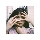 A square portrait of a woman with dark hair posing with her hands over her eyes, wearing a light pink top, set against a blurred urban background with a white border.