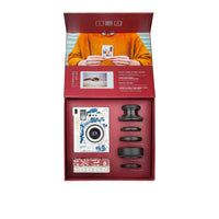 A boxed camera kit displayed open against a light gray background. The set includes a white and blue illustrated instant camera, several black lens attachments, a small red-and-white accessory card, and printed packaging inserts featuring a person in an orange sweater and product text.