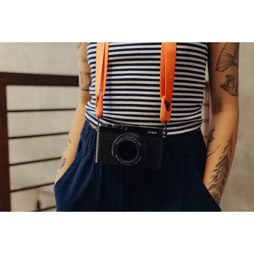 Person wearing a striped shirt and dark pants with an orange strap over their shoulder, holding a black camera.