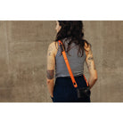 Person with tattoos wearing a striped tank top and carrying a camera with an orange strap against a concrete wall.