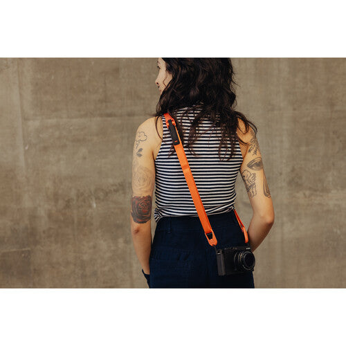Person with tattoos wearing a striped tank top and carrying a camera with an orange strap against a concrete wall.