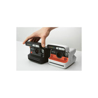 Hand adjusting a black Polaroid Flip instant camera next to a white and orange model on a white surface.