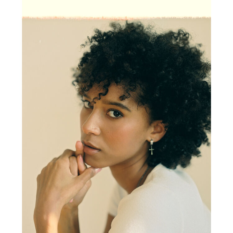 A close-up portrait of a woman with short, curly black hair wearing a white top and a small cross earring, posed against a soft beige background.