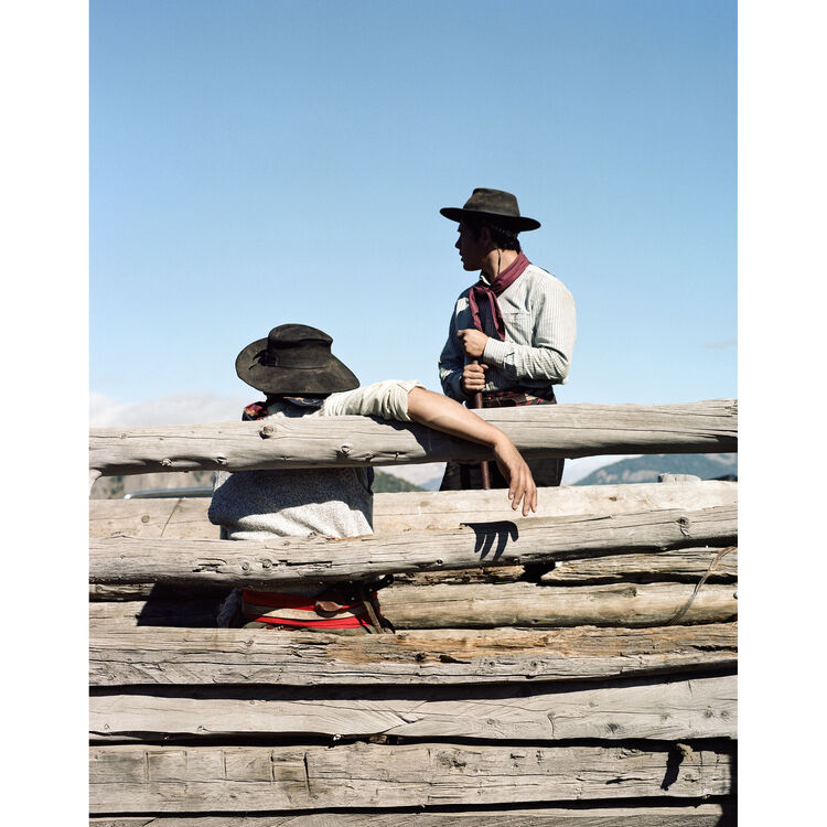 Two people in cowboy hats and western clothing standing and sitting behind rustic wooden logs under a clear blue sky.