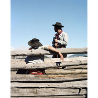 Two people in cowboy hats and western clothing standing and sitting behind rustic wooden logs under a clear blue sky.