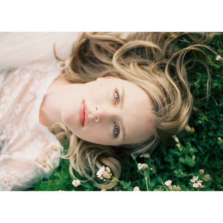 A close-up portrait of a young woman with blonde hair lying in green grass among small white flowers, wearing a pale pink lace garment.
