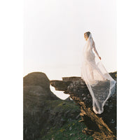 A bride in a long white lace wedding dress with a sheer veil stands on a rocky cliffside outdoors, captured in warm golden-hour light against a pale sky.